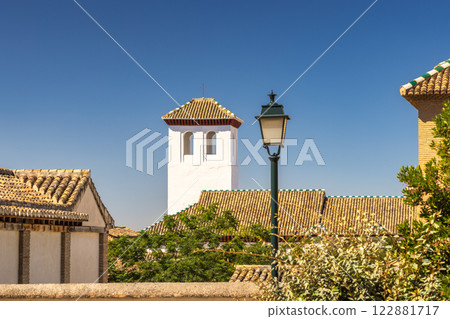 Granada town in Spain. White tower and terracotta rooftops under a clear blue sky, with a lamppost in the foreground. A tranquil, sunny scene. Granada town in Spain. White tower and terracotta rooftops under a clear blue sky, with a lamppost in the foreground. A tranquil, sunny scene. 122881717