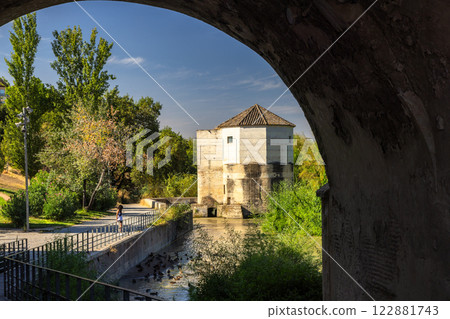 Cordoba town in Spain. Scenic view of a historic stone tower beside a calm waterway, seen through an archway. Cordoba town in Spain. Scenic view of a historic stone tower beside a calm waterway, seen through an archway. 122881743