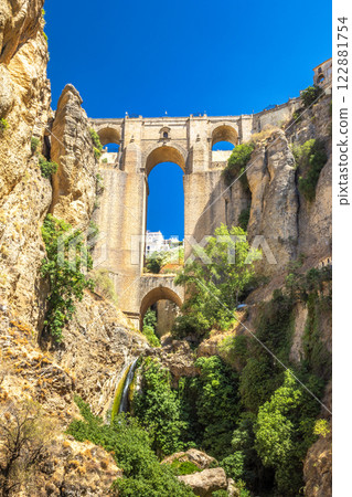 The Puente Nuevo bridge in El Tajo gorge in Ronda town in Spain. Ancient stone bridge spanning a deep gorge, lush vegetation clinging to the rocky walls. A small waterfall cascades down. The Puente Nuevo bridge in El Tajo gorge in Ronda town in Spain. Ancient stone bridge spanning a deep gorge, lush vegetation clinging to the rocky walls. A small waterfall cascades down. 122881754