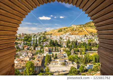 Granada town in Spain from the Alhambra complex. Panoramic view of a city nestled in a hill, seen through an arched window. Sunny day, lush greenery, and traditional architecture. 122881762