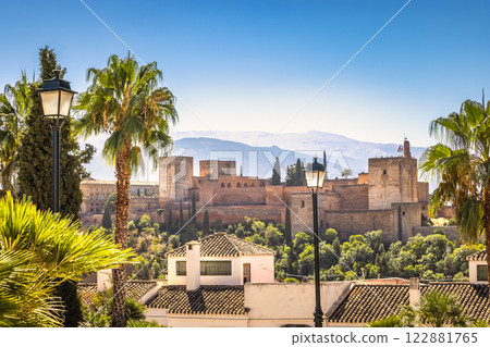 The Alhambra palace from viewpoint in Granada town in Spain. Historic castle nestled in a sunny, palm-tree filled landscape, mountains in the background. 122881765