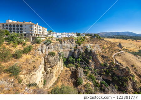 Ronda town in Spain. White village perched on dramatic cliffs, overlooking a vast, dry landscape under a clear blue sky. 122881777