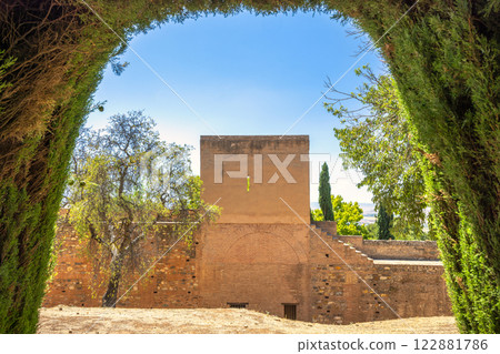 Alhambra complex in Granada town, Andalusia, Spain. Ancient stone wall viewed through a lush green archway, under a clear blue sky. A tranquil, historical scene. Alhambra complex in Granada town, Andalusia, Spain. Ancient stone wall viewed through a lush green archway, under a clear blue sky. A tranquil, historical scene. 122881786