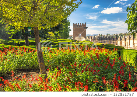 Alhambra complex in Granada town, Andalusia, Spain. Vibrant flowers bloom in a meticulously manicured garden, with a historic stone wall and tower in the background. A sunny day. Alhambra complex in Granada town, Andalusia, Spain. Vibrant flowers bloom in a meticulously manicured garden, with a historic stone wall and tower in the background. A sunny day. 122881787