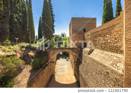 Aqueduct of the Acequia Real in the Alhambra complex, Granada town, Andalusia, Spain. Ancient stone archway and fortress walls under a clear blue sky. Tall cypress trees frame the scene. Aqueduct of the Acequia Real in the Alhambra complex, Granada town, Andalusia, Spain. Ancient stone archway and fortress walls under a clear blue sky. Tall cypress trees frame the scene. 122881790