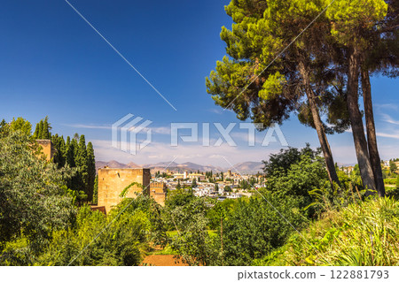 Granada town in Spain. Panoramic view of a historic city nestled in a valley, seen from a lush garden with tall trees. Blue skies and mountains complete the scene. 122881793