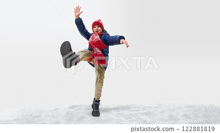 Boy dressed blue winter jacket, with red scarf jumping on snow, stretching arms wide, enjoying playful winter moment against white studio background. 122881819