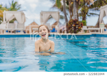 A young boy joyfully swims in a tropical pool surrounded by lush greenery. The vibrant scene captures the essence of summer fun, adventure, and carefree childhood moments in a refreshing outdoor 122881844