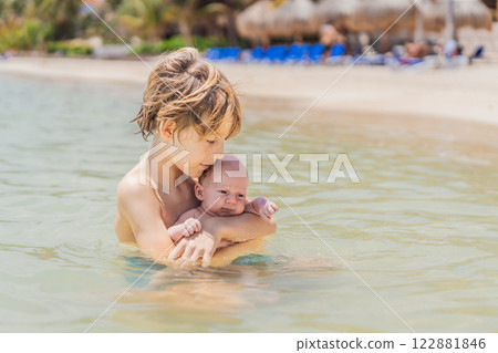 An older brother gently holds his baby sibling while swimming in the sea, sharing a joyful and heartwarming moment of family bonding and summer fun in the water. A perfect blend of love, care, and 122881846
