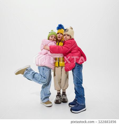 Three children, dressed warm winter jackets, hugging each other with smiles, showing friendship and joy of childhood against white studio background. Three children, dressed warm winter jackets, hugging each other with smiles, showing friendship and joy of childhood against white studio background. 122881856