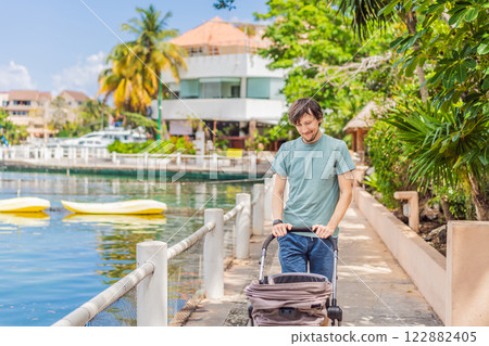A young father walks along the scenic waterfront, pushing a baby stroller with her child inside. A peaceful and heartwarming moment of motherhood, relaxation, and outdoor bonding by the sea 122882405
