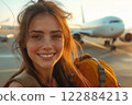 A cheerful young woman stands at the airport, holding her backpack, with a commercial airplane parked behind her. The warm light of sunset highlights her joyful expression. 122884213