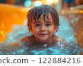 A young boy with wet hair and bright eyes smiles while swimming in a vibrant indoor pool. The setting is lively, filled with warm lights reflecting off the water. 122884216