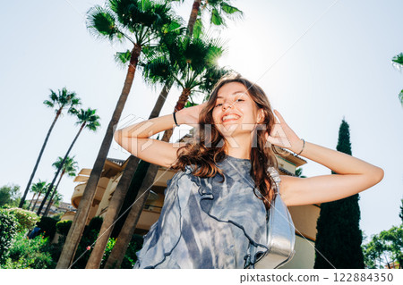 A young woman in a flowing gray dress smiles brightly, walking among palm trees with a modern A young woman in a flowing gray dress smiles brightly, walking among palm trees with a modern 122884350