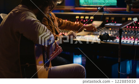 Creative musician singing guitar in professional studio control room, recording his tracks with producer. Singer songwriter composing a new song for his album, editing tunes on console. Camera A. 122885010