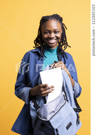 Portrait of upbeat BIPOC young girl browsing through backpack, isolated over studio background. Radiant college student taking out textbooks out of rucksack used during commuting to university 122885103