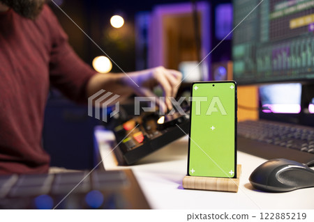 Sound engineer uses controls and amplifier on mixing console to create beautiful sound effects and adjust volume levels on tracks. Producer using greenscreen display on phone, editing soundtracks. 122885219
