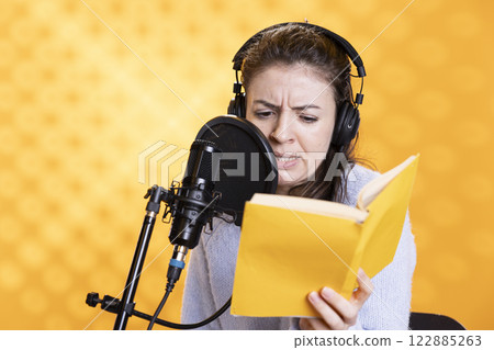 Narrator frowning while reading book, recording audiobook using microphone, playing angry character, studio background. Woman producing digital recording of novel, interpreting furious antagonist Narrator frowning while reading book, recording audiobook using microphone, playing angry character, studio background. Woman producing digital recording of novel, interpreting furious antagonist 122885263