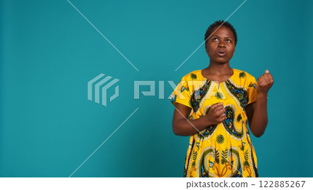 Optimistic glamorous girl praying for some good luck and fortune, feeling positive and hopeful against blue background. Young woman in traditional tribal attire in studio. Camera B. 122885267