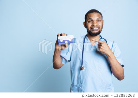 Portrait of doctor in scrubs recommending vitamins and supplements for wellness, holding medicine box for patients in studio. Hospital employee pointing at tablets for chronic disease. 122885268