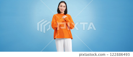 Care and love. Asian woman holds hands on heart and smiles, looks with tenderness and warmth at camera, stands over blue background Care and love. Asian woman holds hands on heart and smiles, looks with tenderness and warmth at camera, stands over blue background 122886493