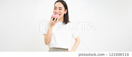 Close up of smiling pleased asian woman, eats glazed pink doughnut, enjoys eating tasty donnut, white studio background 122886518