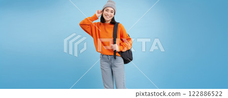 Cute smiling asian girl puts on warm hat to go outside, walks with backpack in orange sweater, blue background Cute smiling asian girl puts on warm hat to go outside, walks with backpack in orange sweater, blue background 122886522