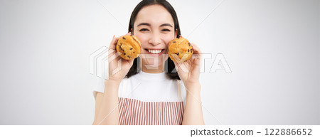 Image of korean woman enjoys eating baked pastry, showing two tasty cupcakes near face and smiles, white background Image of korean woman enjoys eating baked pastry, showing two tasty cupcakes near face and smiles, white background 122886652