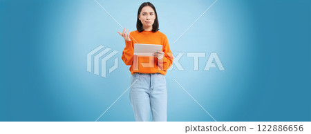 Woman with digital tablet, looks confused, cant understand smth on her gadget, stands puzzled against blue background 122886656
