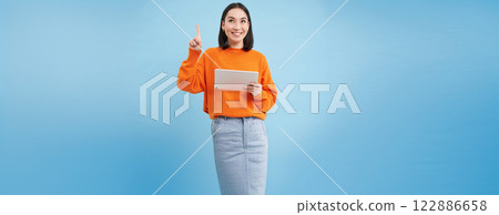 Beautiful young korean woman with tablet, pointing finger up and looking on top advertisement, showing banner, standing over blue background. Online shopping concept 122886658
