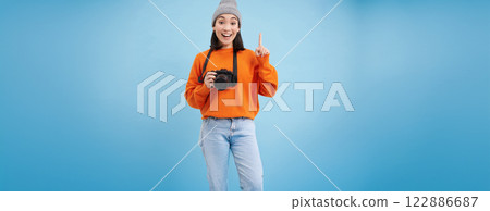 Surprised asian girl in hat, holds digital camera, looks amazed and impressed, poses over blue studio background with space for banner Surprised asian girl in hat, holds digital camera, looks amazed and impressed, poses over blue studio background with space for banner 122886687