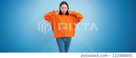 Cant listen. Fed up asian woman shuts her ears with fingers and closes eyes from loud noise, stands in orange sweatshirt over blue background 122886691