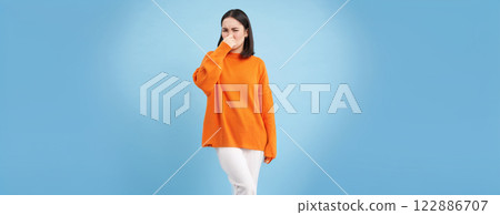 Something stinks. Young asian woman shuts her nose with fingers from bad smell, wears orange sweatshirt, stands over blue background 122886707
