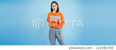 Woman with digital tablet thinking, looking aside with thoughtful face expression, standing over blue background Woman with digital tablet thinking, looking aside with thoughtful face expression, standing over blue background 122886708
