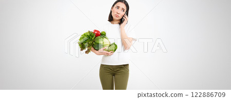 Portrait of sad, unhappy young woman, making phone call, talking with someone and holding vegetables, white background 122886709