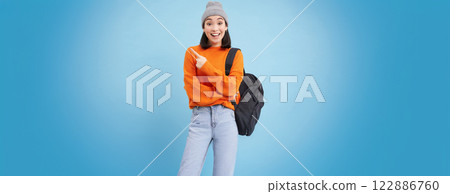 Enthusiastic asian woman points left, shows promo offer, holds backpack, stands over blue background 122886760