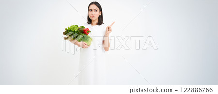 Portrait of pensive asian woman, holding vegetables, pointing right with thinking face, white background Portrait of pensive asian woman, holding vegetables, pointing right with thinking face, white background 122886766