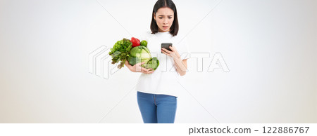 Portrait of asian woman looks worries at smartphone screen, holds vegetables, orders food online, white background Portrait of asian woman looks worries at smartphone screen, holds vegetables, orders food online, white background 122886767