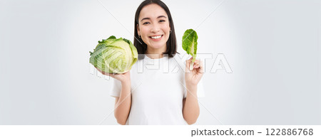 Close up portrait of asian woman, biting lettuce, eating green cabbage and smiling, white background 122886768