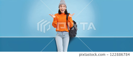 Happy asian woman in warm hat, claps hands from surprise, laughs and looks amazed, excited by smth, stands over blue background 122886784