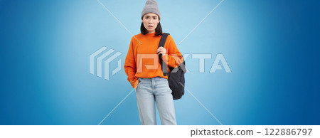 Young asian woman in hat, holds backpack, sulks and looks confused, perplexed emotion, stands over blue background 122886797