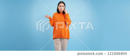 Sad and confused asia girl shrugs, talks on cellphone, answers telephone call with puzzled face, stands over blue background Sad and confused asia girl shrugs, talks on cellphone, answers telephone call with puzzled face, stands over blue background 122886804
