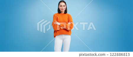 Young asian woman looks with confidence, cross arms on chest and smiles at camera, stands in orange sweatshirt, blue background Young asian woman looks with confidence, cross arms on chest and smiles at camera, stands in orange sweatshirt, blue background 122886813