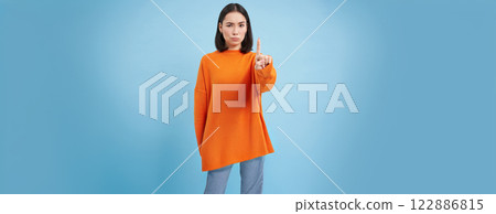 Rule number one. Serious korean girl shows finger, shakes forefinger, disapproves smth, prohibits, stands over blue background Rule number one. Serious korean girl shows finger, shakes forefinger, disapproves smth, prohibits, stands over blue background 122886815