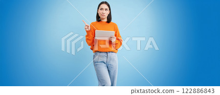Portrait of korean woman with digital tablet, pointing at upper left corner with confused, frowning face, standing over blue background Portrait of korean woman with digital tablet, pointing at upper left corner with confused, frowning face, standing over blue background 122886843