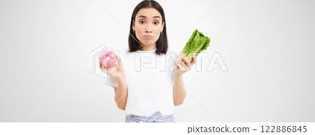 Healthy lifestyle concept. Portrait of sad complicated asian woman, shows doughnut and cabbage, decides between vegetables and diet or delicious junk food, white background 122886845