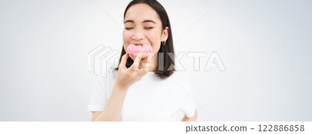 Food and eating out. Young korean woman looks, takes bite of delicious pink glazed doughnut, white background Food and eating out. Young korean woman looks, takes bite of delicious pink glazed doughnut, white background 122886858