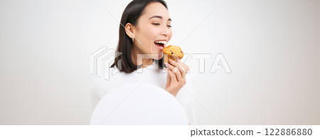 Close up of happy korean woman eating cupcake, delicious dessert, smiling from pleasure, white background 122886880