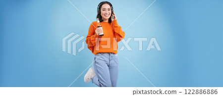 Beautiful asian woman listens music in headphones, drinks coke from bottle and smiles at camera, blue background 122886886