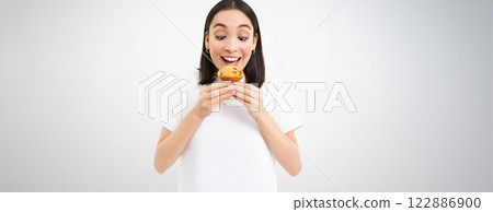 Close up portrait of asian woman likes pastry, bites tasty cupcake with happy unbothered smile, white background 122886900